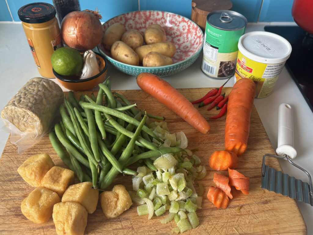 Ingredients layout on a wooden board for Thai yellow curry, featuring fresh green beans, chopped celery, diced carrots, and new potatoes.